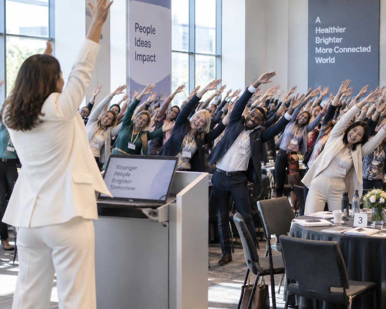 A leader at a podium prompting a large audience to raise arms and lean together